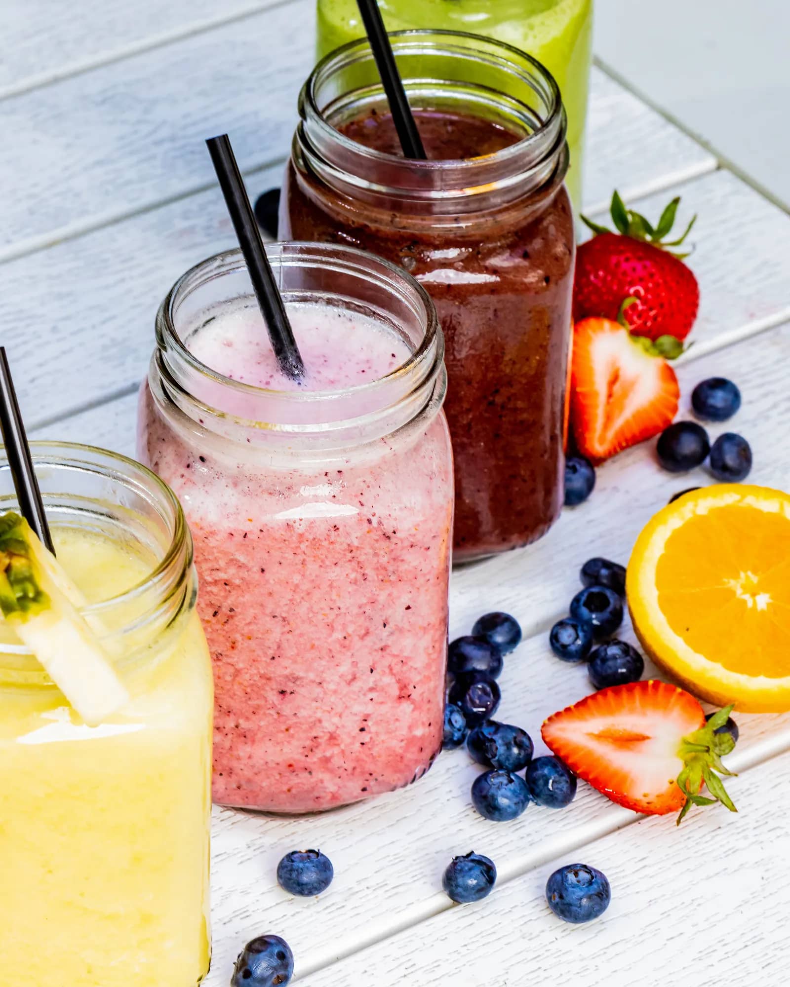 Assorted fresh smoothies and juice in mason jars surrounded by strawberries, blueberries and orange