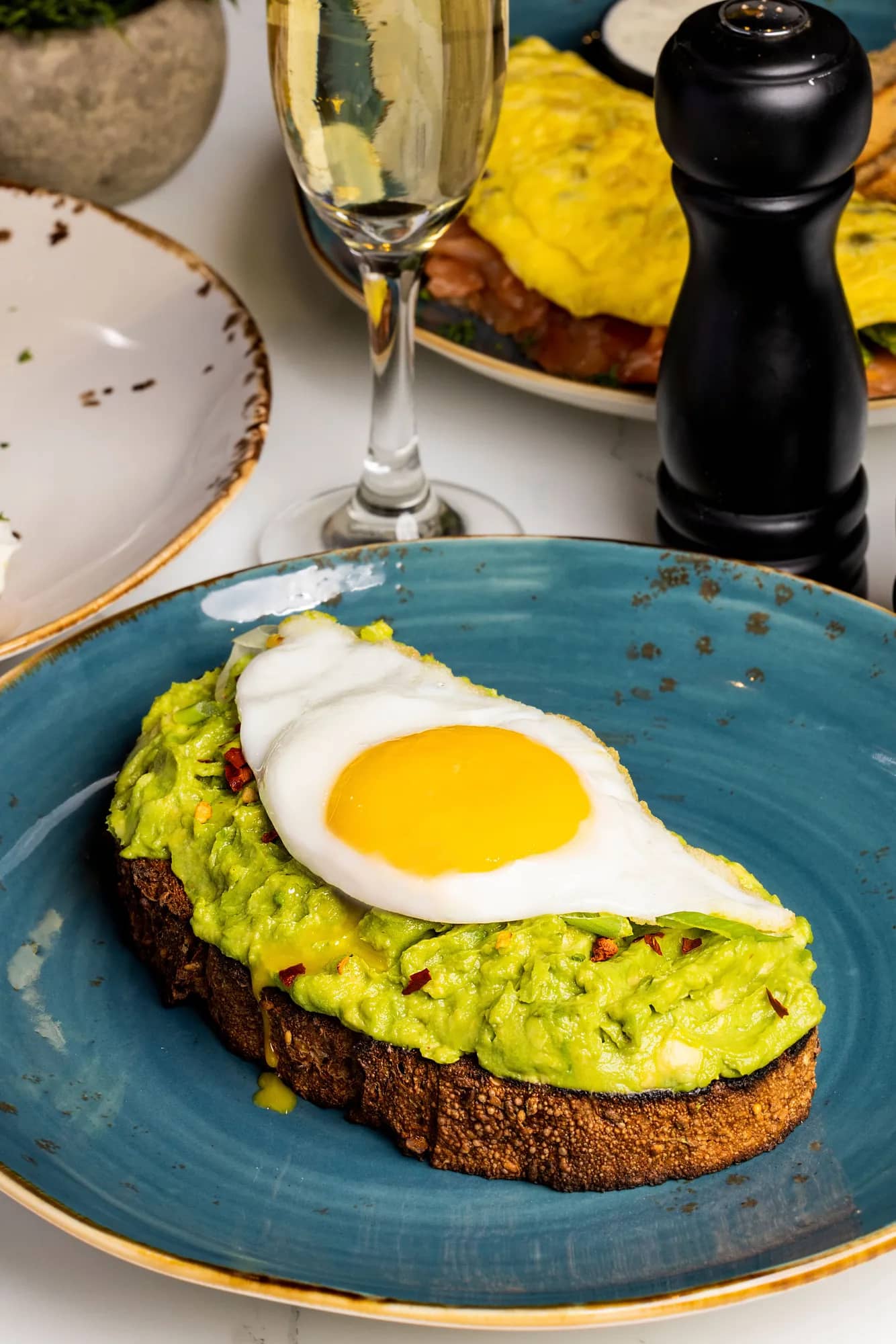 Avocado toast topped with a fried egg on a blue plate, with a glass of sparkling wine nearby