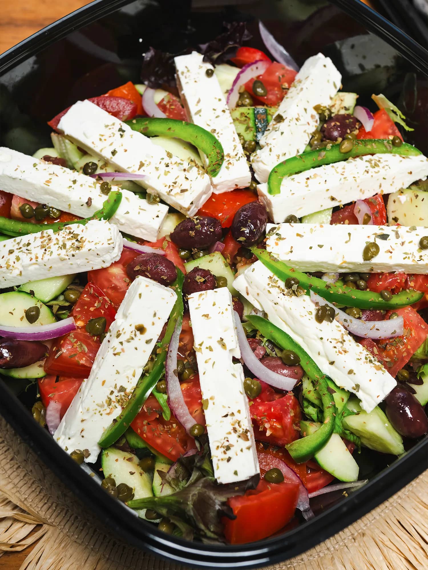 Greek salad in a black catering container with feta slices, tomatoes, cucumber, peppers, olives and oregano